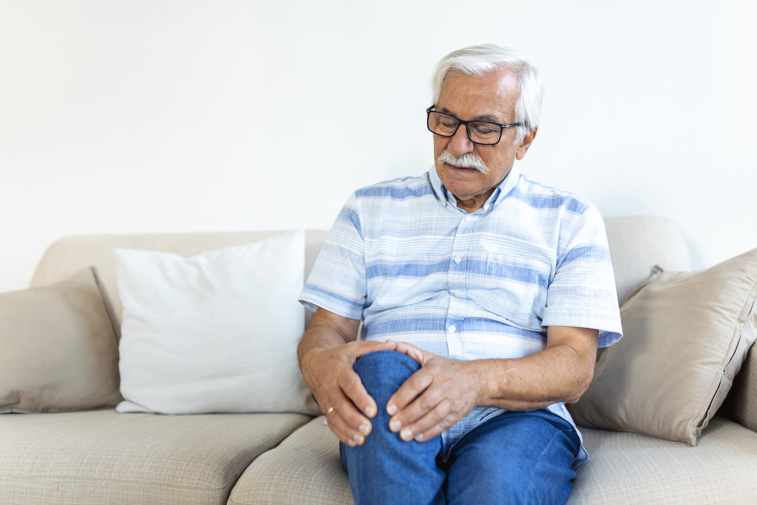 Elderly man sitting on a sofa at home and touching his painful knee. people, health care and problem concept – unhappy senior man suffering from knee ache at home